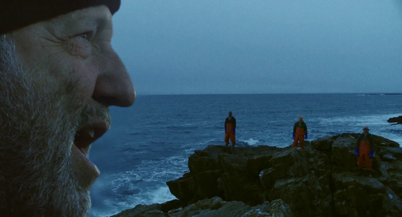 Blow The Man Down (2019) movie still: wide — A dark rocky shoreline meets a cold, deep blue ocean at twilight. Three men in orange waterproof work…