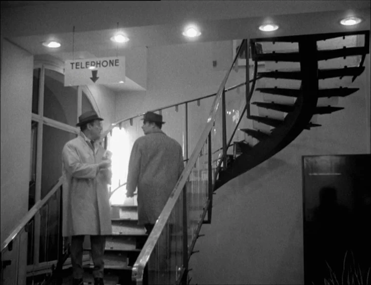 Alphaville (1965) movie still: fullbody — A modern building interior features a sweeping spiral staircase with a metal railing. Two men wearing hats…