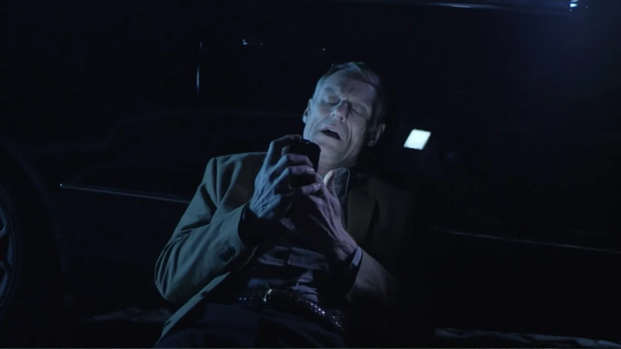 Entre las sombras (2025) movie still: medium — The interior of a dark car is illuminated by a small handheld electronic device. An older man sits in…