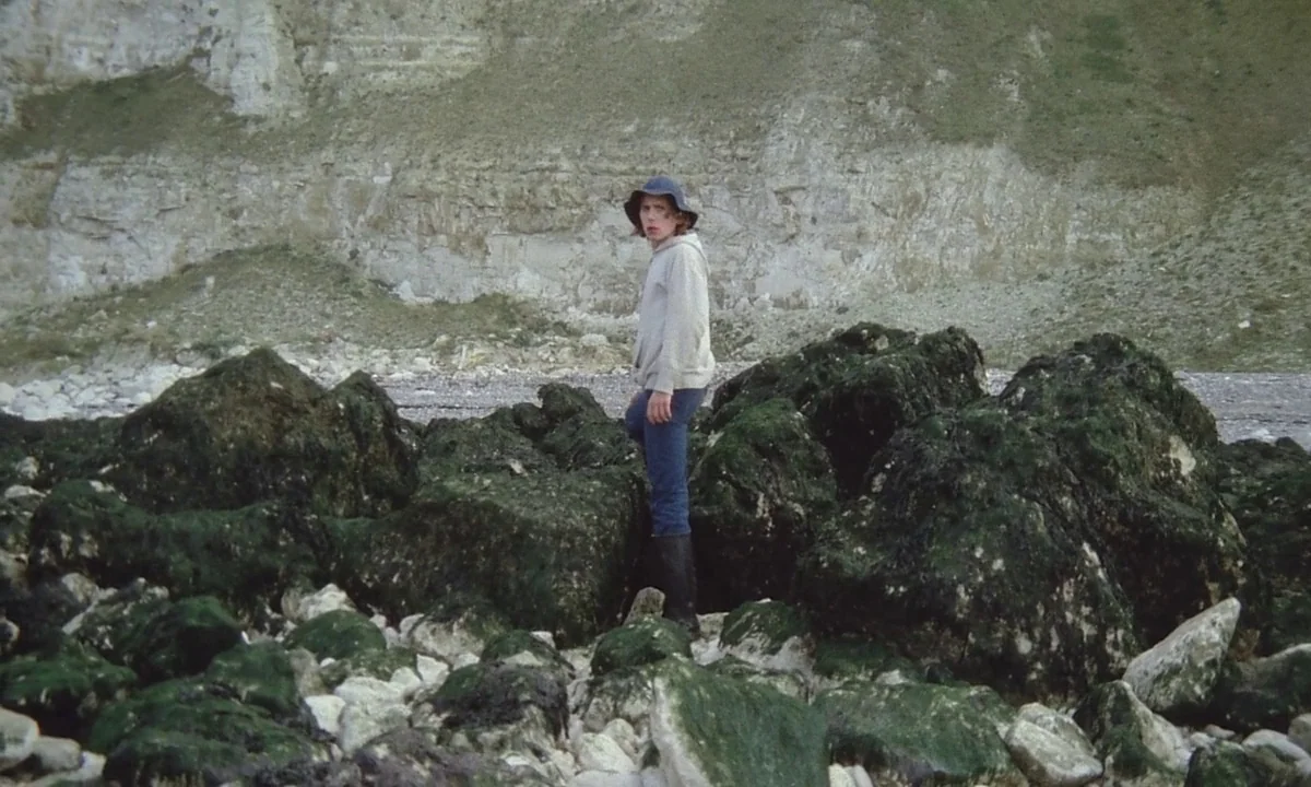 Immoral Tales (1974) movie still: fullbody — A rocky beach shoreline sits at the base of a high white chalk cliff face. A young man stands on the…