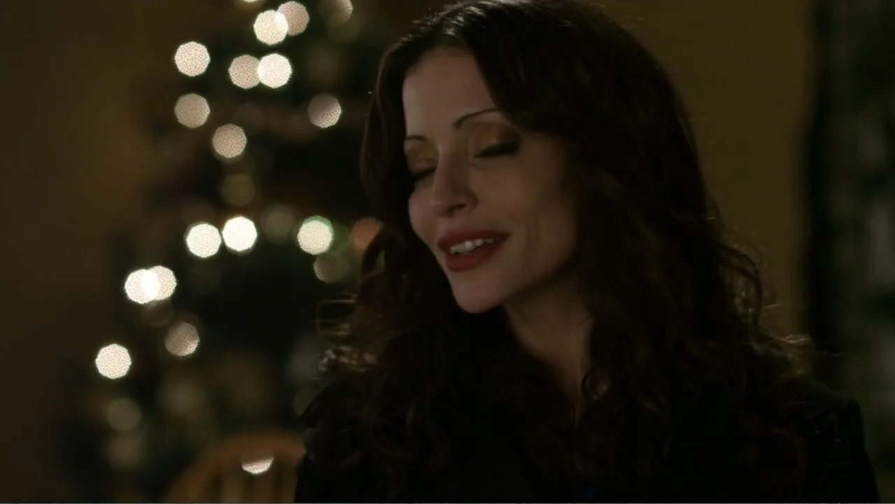 It's Christmas, Carol! (2012) movie still: closeup — An interior space features a woman in the foreground with blurred holiday lights behind. The…
