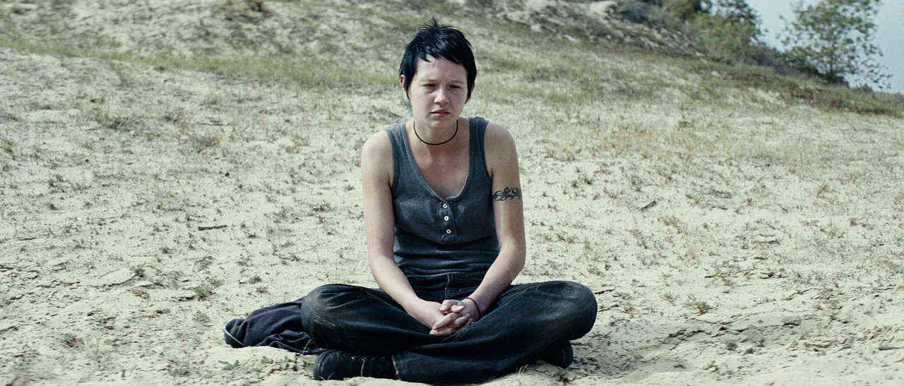 Outside Satan (2011) movie still: fullbody — A desolate sandy landscape with sparse vegetation under an overcast sky. A young woman sits on the…
