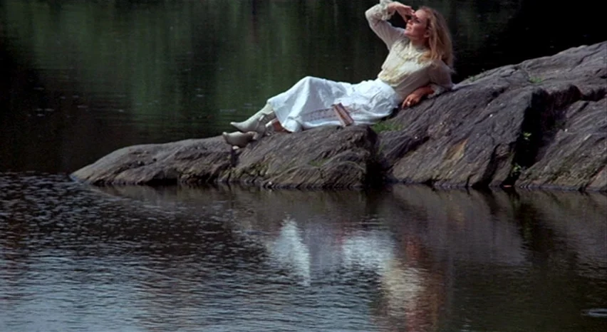 A Safe Place (1971) movie still: wide — A woman reclines on a dark rock surrounded by dark water. She shields her eyes from the sunlight while…