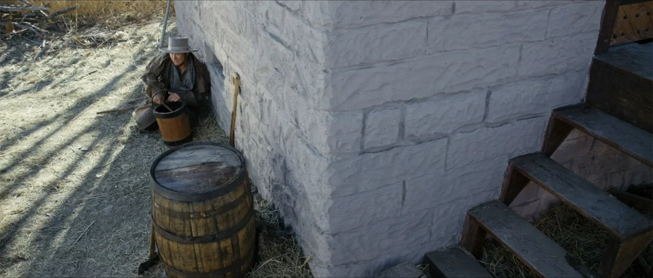 Out of Liberty (2019) movie still: wide — An exterior homestead area features a rough-hewn stone wall and wooden stairs. A man in a duster coat sits…