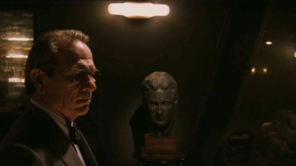 A Prairie Home Companion movie still: medium — A dark study contains a sculpted bust on a stand next to a man. The man in a grey suit gazes past the…