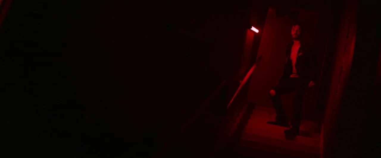 VFW movie still: wide — A dark stairwell is lit primarily by a red neon tube mounted on the wall. A man stands on the stairs looking up toward the…