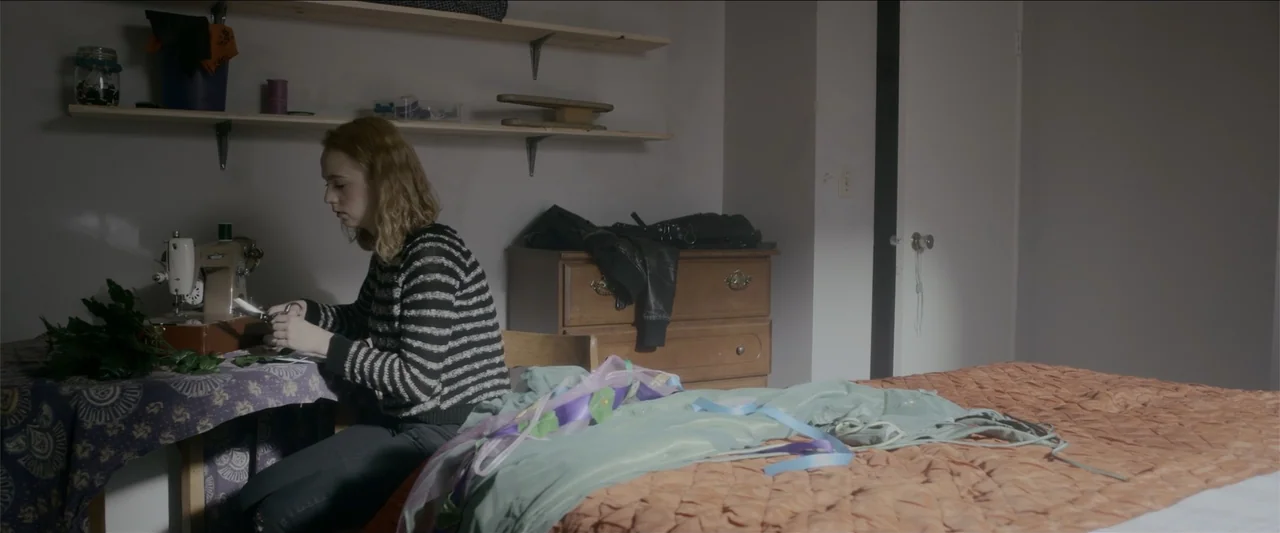 The Hollow Child (2018) movie still: medium — A spare bedroom contains a wooden dresser, a sewing machine, and a bed. A young woman sits at the desk…