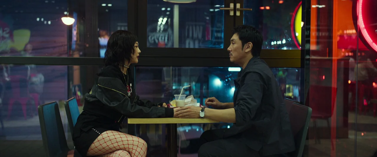 Believer (2018) movie still: medium — A window-seat booth inside a brightly lit fast-food eatery at night. A man and a young woman sit face-to-face,…