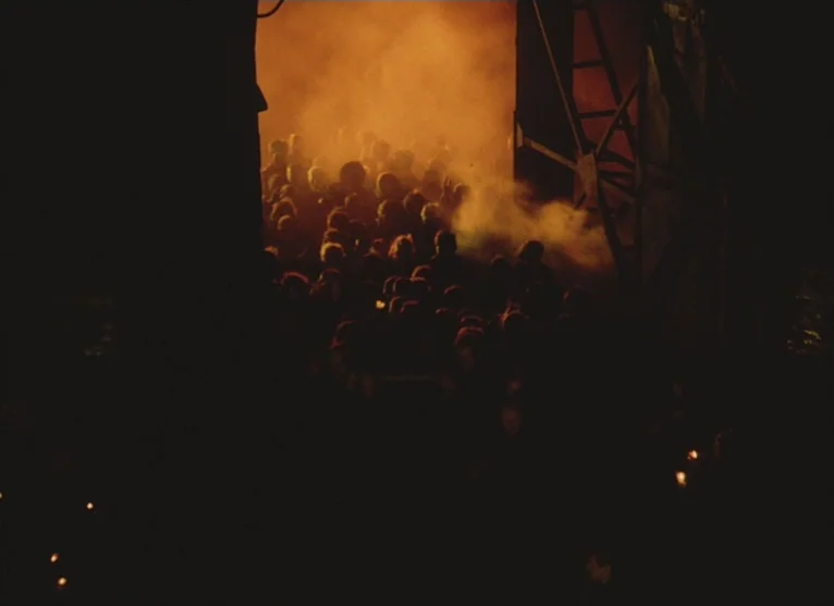 A Visitor to a Museum (1989) movie still: wide — A dark, industrial doorway frames a crowd moving through dense, orange-lit haze. The figures are…