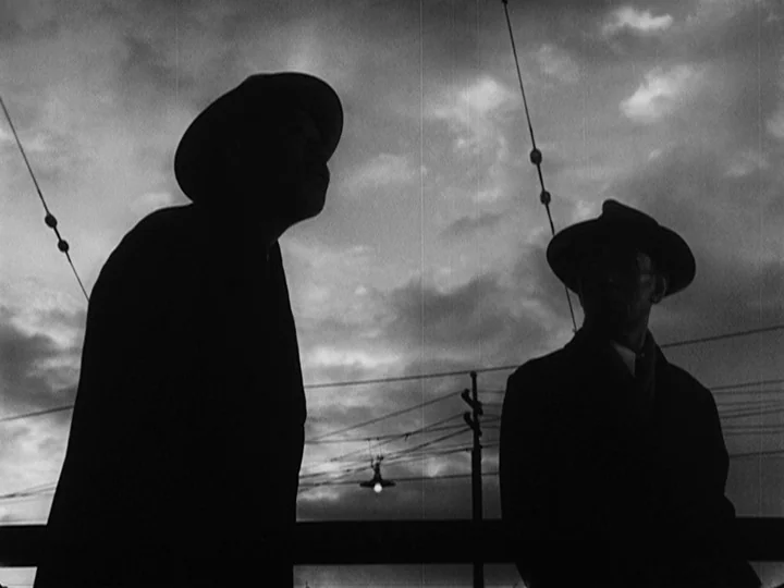 Ikiru (1952) movie still: medium — Two men in hats and coats stand on an elevated outdoor platform at twilight. Overhead power lines crisscross…