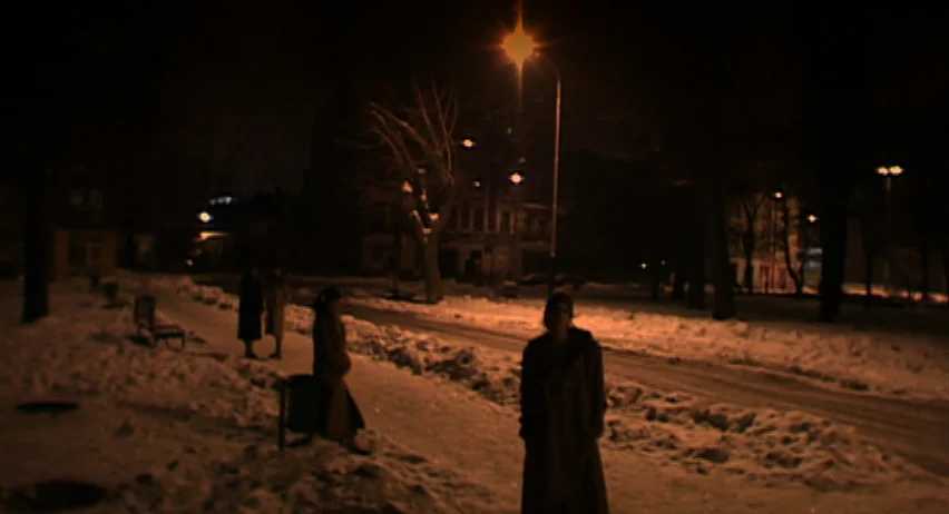 Inland Empire (2006) movie still: wide — A snow-covered public space at night is illuminated by a lone overhead street lamp. Several people stand at…