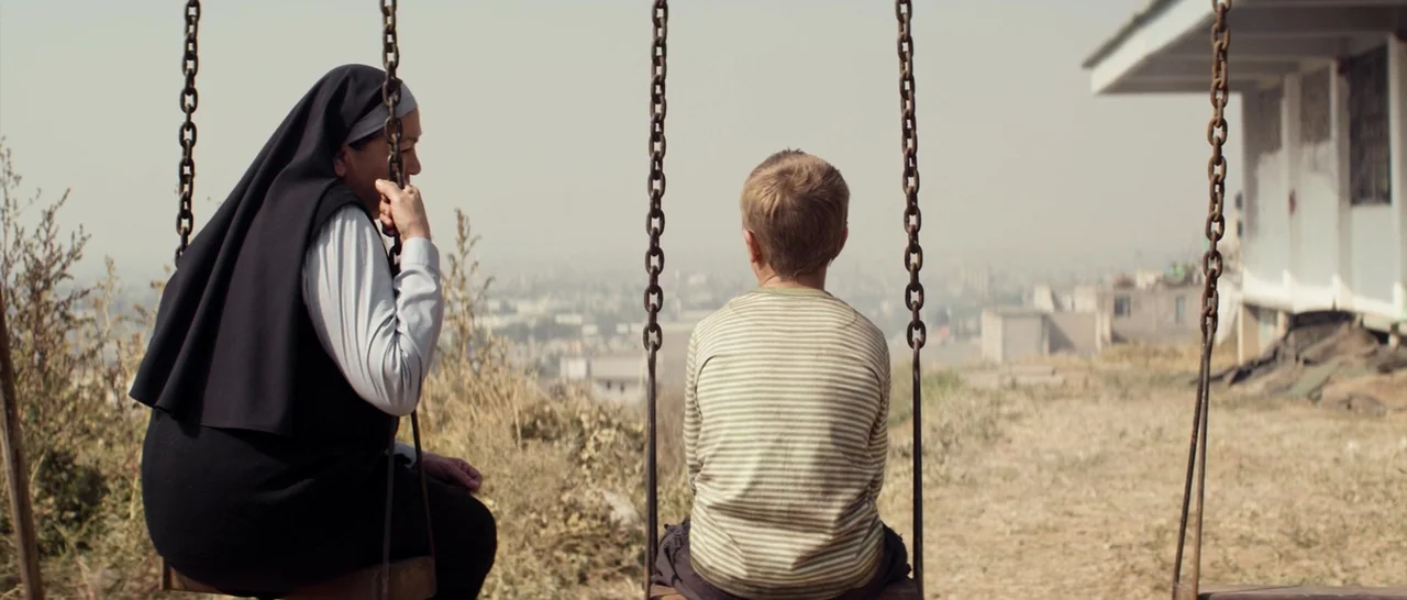 Elysium (2013) movie still: medium — A rural playground on a hillside overlooking a hazy, sprawling desert city. A nun and a young boy sit on…