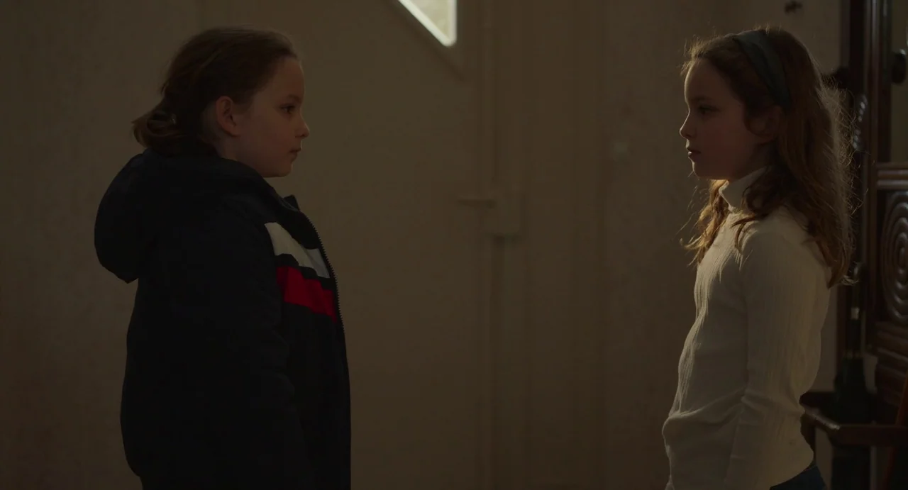 Petite Maman (2021) movie still: medium — A dimly lit indoor space with textured walls and a wooden structure nearby. Two young girls stand…