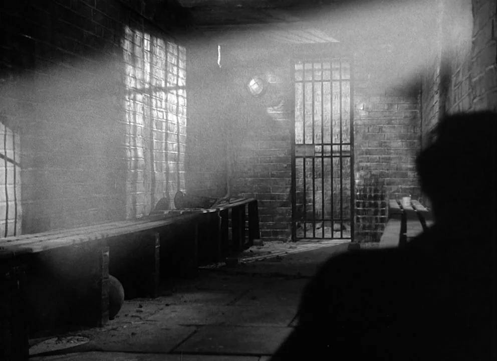 Odd Man Out (1947) movie still: medium — A stone holding cell features heavy brick walls and a metal grate door. A person sits on a wooden bench,…