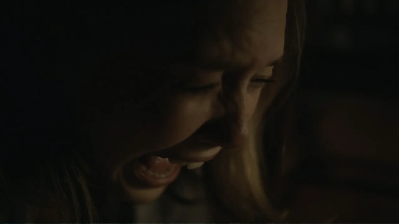 Silent House (2011) movie still: closeup — A dimly lit interior space with the face of a woman emerging from deep shadows. The woman has her mouth…