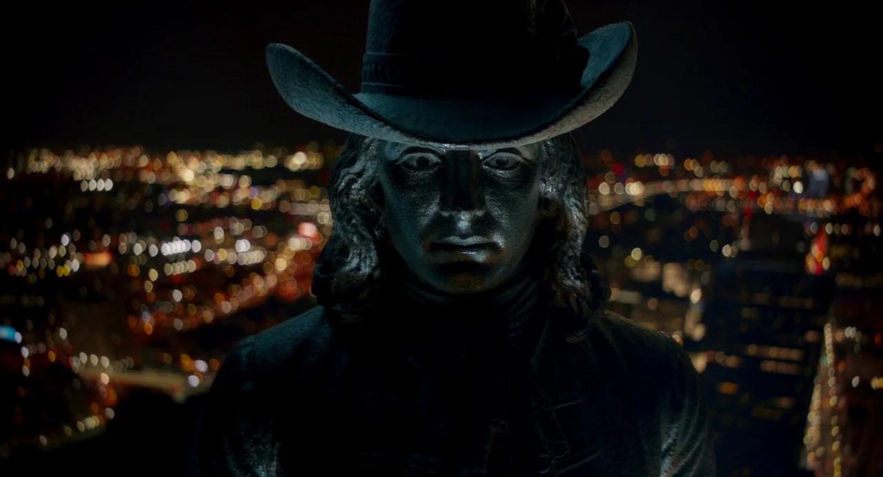 Drive-Away Dykes (2024) movie still: closeup — A bronze statue of Benjamin Franklin stands on a high urban rooftop at night. The city landscape…