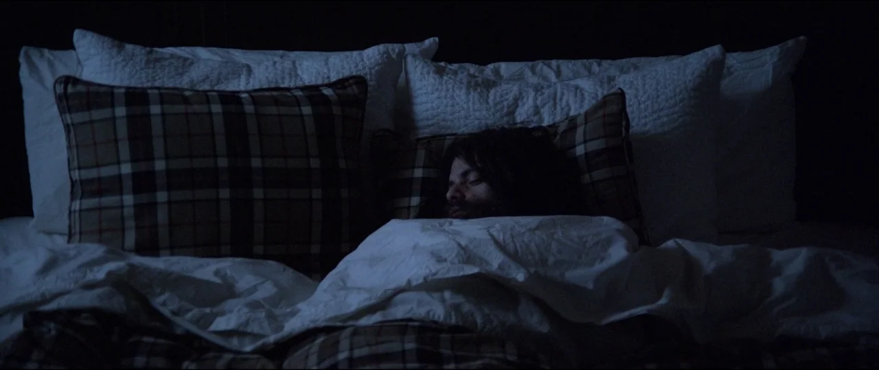 Buster’s Mal Heart (2016) movie still: medium — A dark bedroom features a bed with white and plaid patterned pillows and bedding. A man with long…