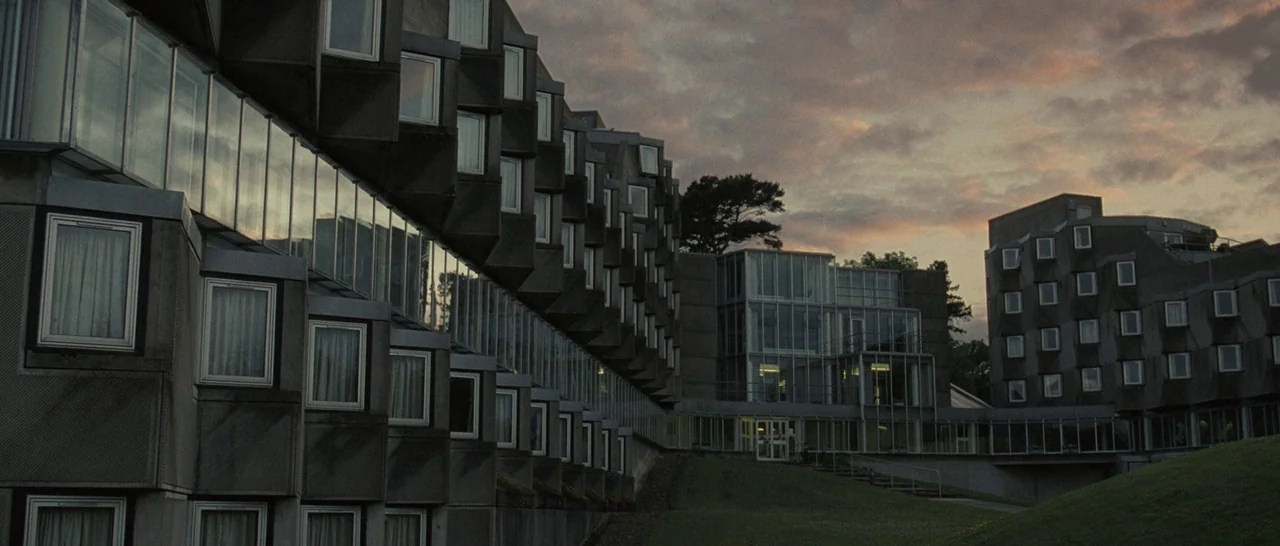 Never Let Me Go (2010) movie still: establishing — A complex of concrete brutalist buildings with repeating angular windows sits on a grassy slope.…