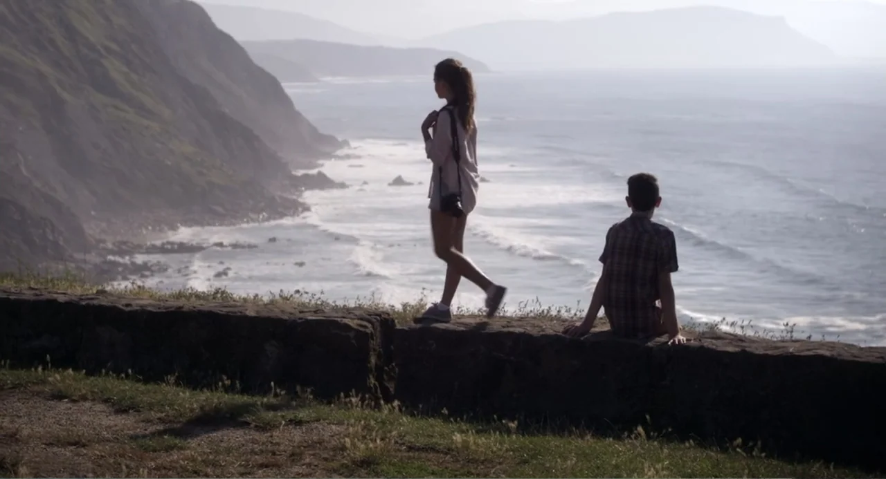 Newton's Third Law (2017) movie still: fullbody — A stone barrier separates a grassy clifftop from a wide ocean view with cliffs. A young woman…