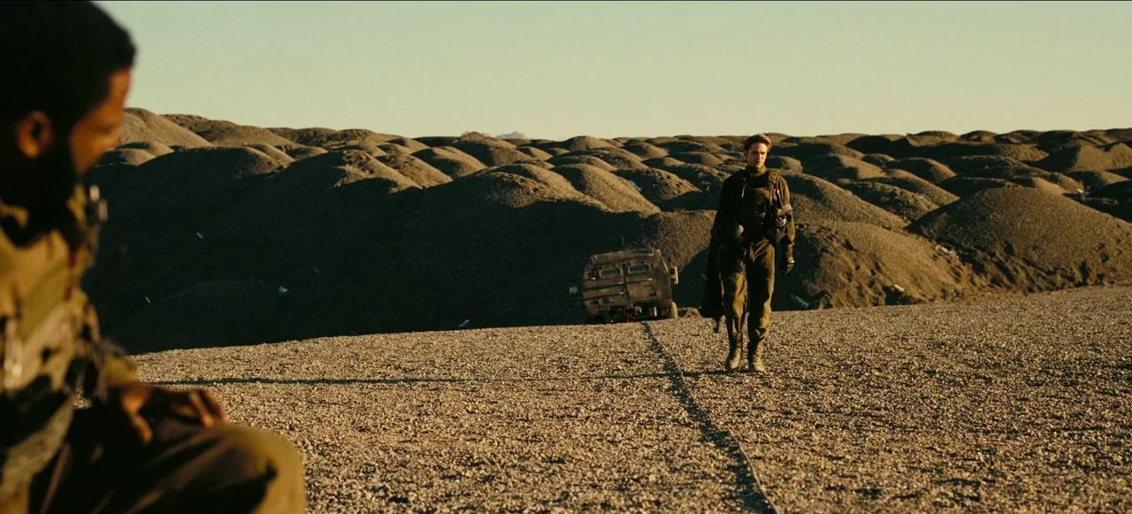 Tenet (2020) movie still: wide — An expanse of rolling, dark gravel hills stretches to the horizon under clear skies. A man in olive-drab tactical…