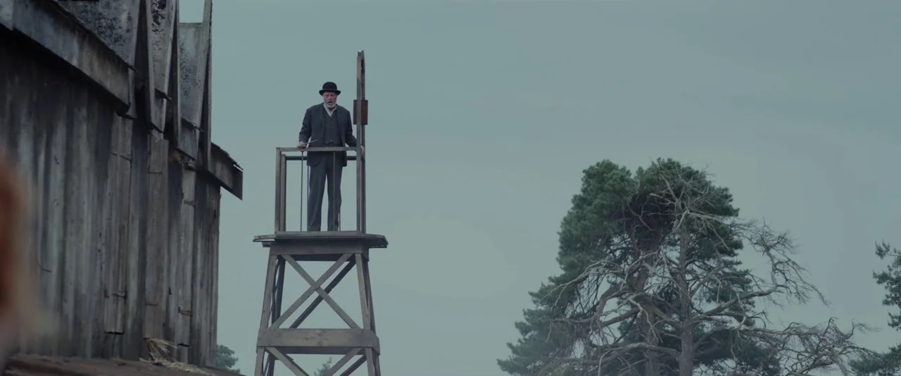 Apostle (2018) movie still: wide — A wooden watchtower rises above a desolate outdoor area with overcast skies and trees. A man in a dark suit…