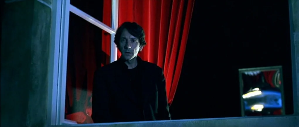 Mortal Transfer (2001) movie still: medium — A man stands behind a window at night flanked by intense red curtains. The dark frame is punctuated by…