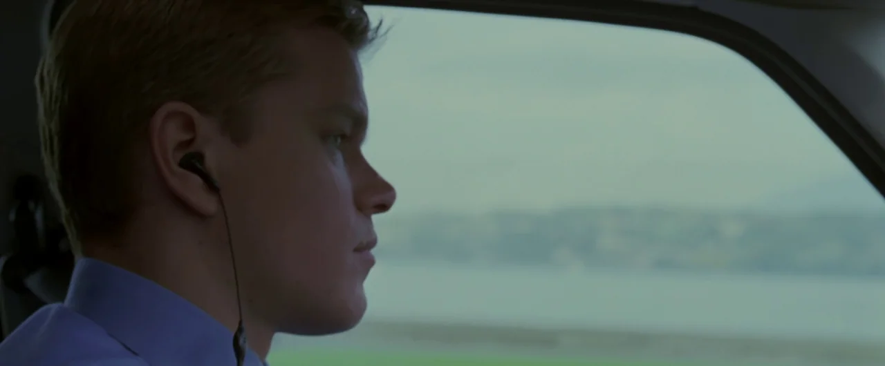 Syriana (2005) movie still: closeup — A side-profile view of a man inside a car against a blurred landscape. The man wears white earphones and gazes…