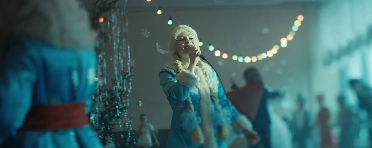 Petrov’s Flu (2021) movie still: medium — A festive indoor space is decorated with tinsel and strings of colored party lights. A woman in a blue…
