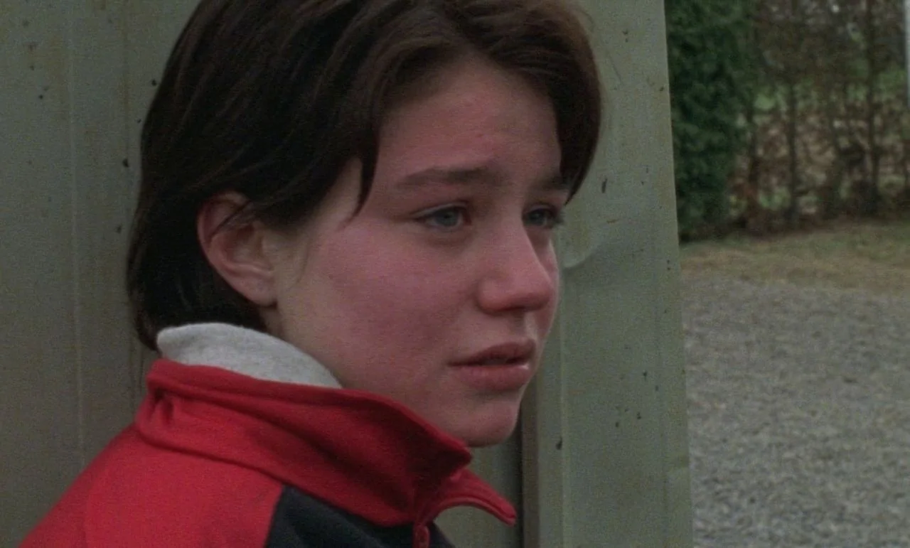Rosetta (1999) movie still: closeup — A person leans against a vertical structure in an outdoor, gravel-filled area. The teenager looks toward the…