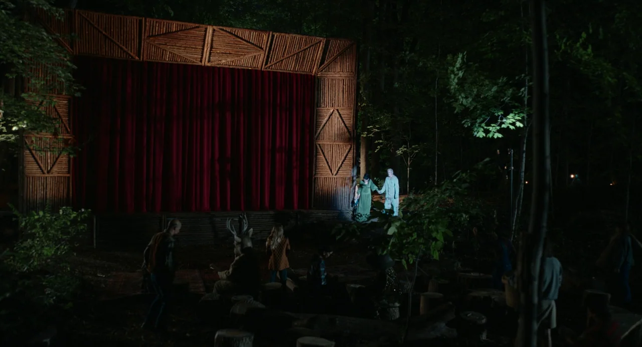 Beau Is Afraid (2023) movie still: wide — A rustic wooden outdoor stage sits within a dark forest at night. Two performers holding hands stand near…