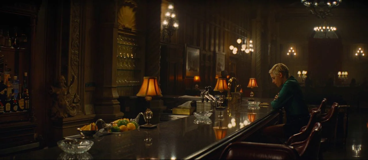 Seberg (2019) movie still: wide — A dark, wood-paneled upscale hotel bar features empty red leather stools and dim lighting. A woman with short…
