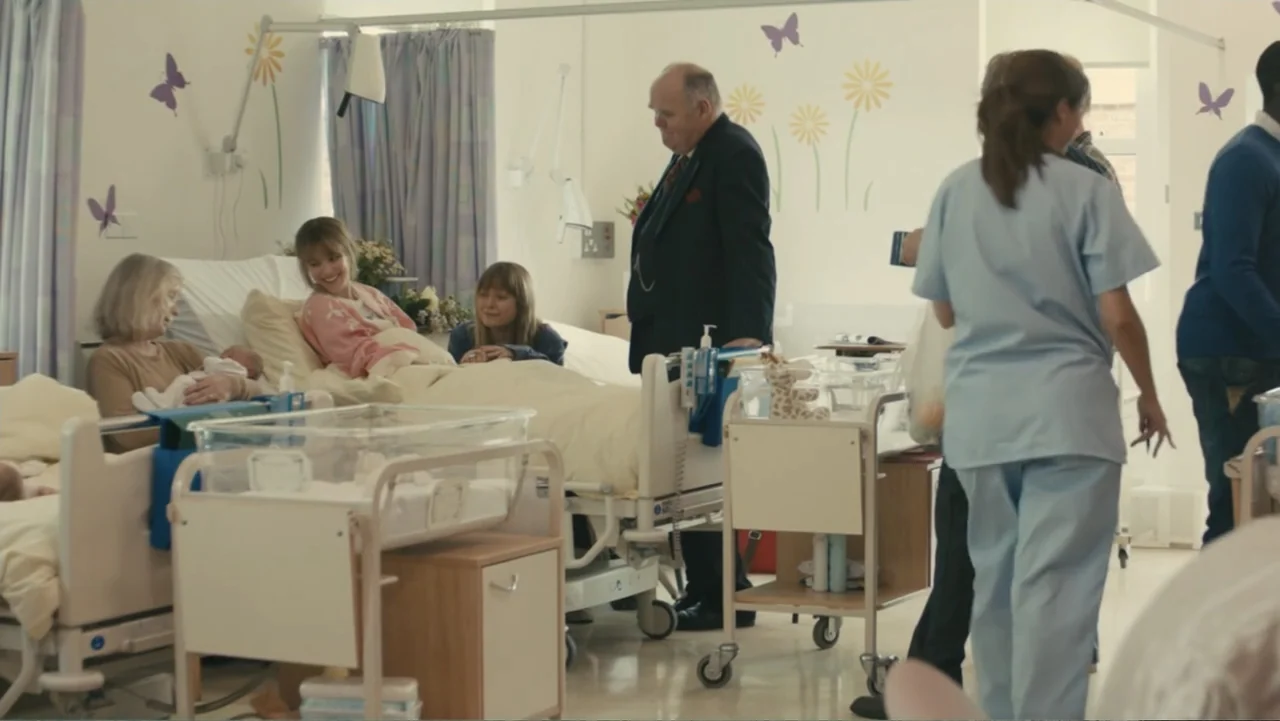 About Time (2013) movie still: wide — A maternity ward features white walls decorated with butterfly and flower wall decals. A woman sits in a bed…