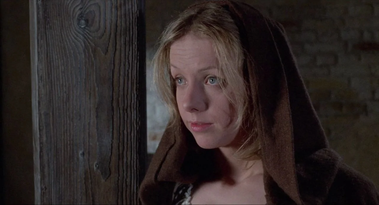 The Blood on Satan’s Claw (1971) movie still: closeup — An interior space features a rough wooden vertical beam and a brick wall. A woman in a dark…