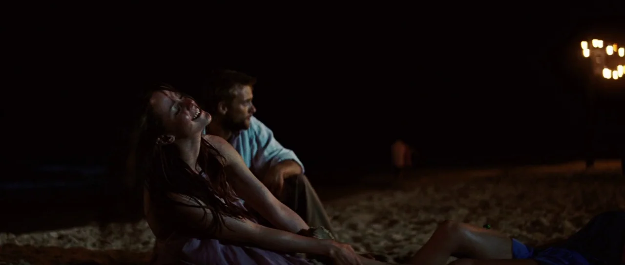 The Ruins (2008) movie still: medium — A dark beach at night with sand illuminated by a distant artificial light. A woman sits on the sand smiling…