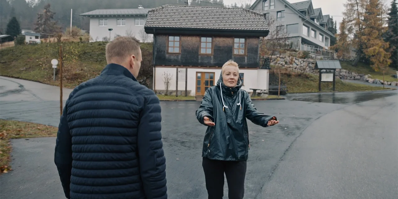 Navalny (2022) movie still: medium — A paved area sits in front of a multi-story rustic building during daytime. A woman speaks to a man who faces…