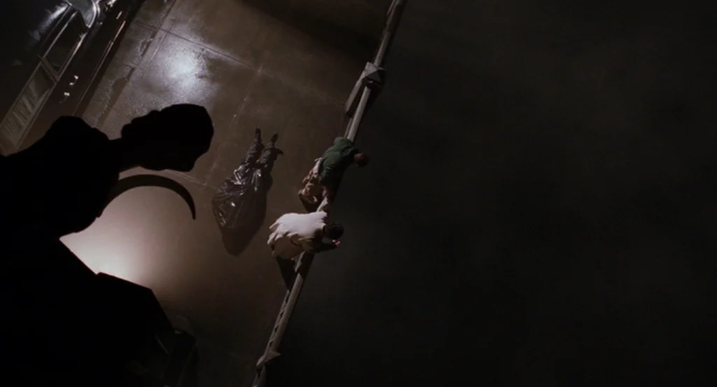 The Ladykillers (2004) movie still: wide — The concrete surface of a high structure at night shows long, sharp cast shadows. Two individuals stand…