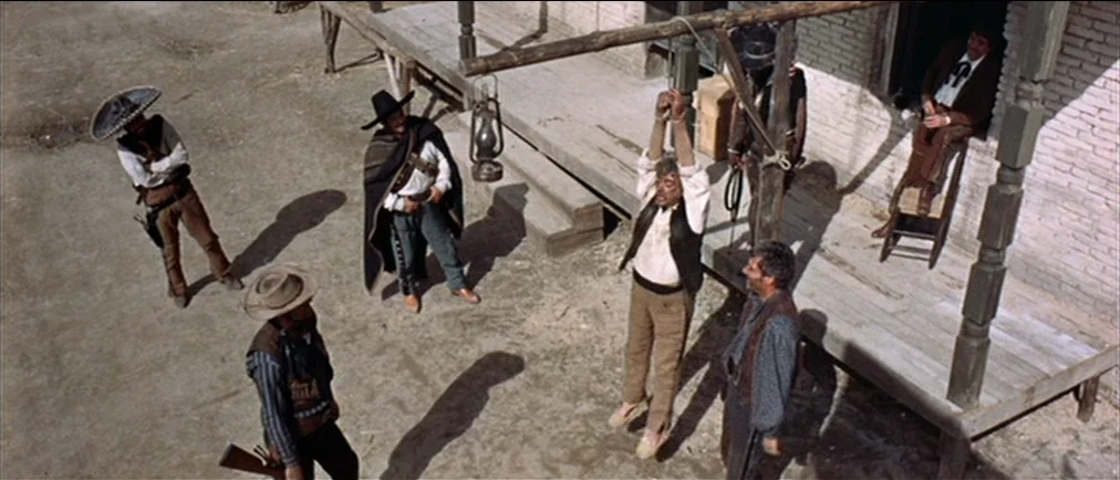 Fistful Of Dollars (1964) movie still: wide — A dusty outdoor space with a wooden structure features a man hanging by his wrists. Several armed men…
