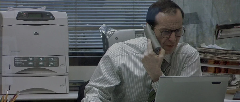 A Mighty Heart (2007) movie still: medium — A workspace with a stack of paper documents and a large HP printer nearby. A man sits in front of his…