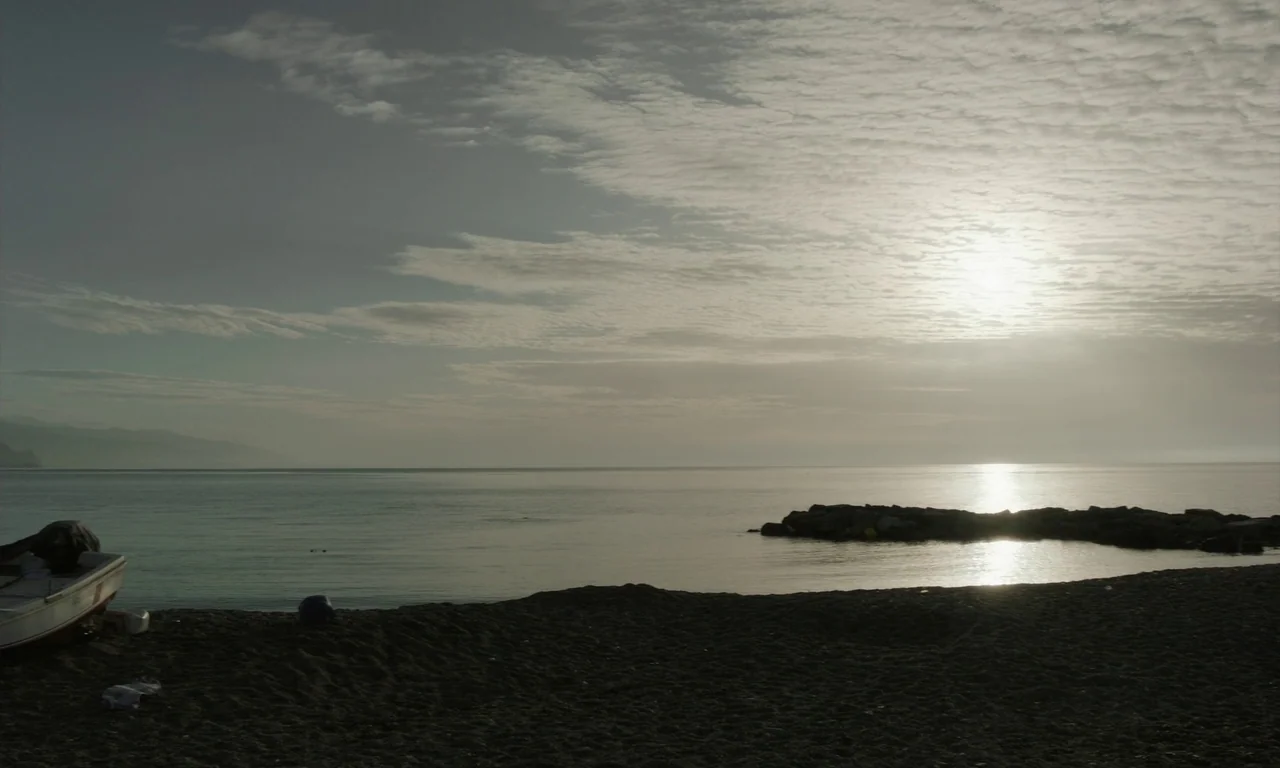 Close Your Eyes (2023) movie still: wide — A calm shoreline features a dark sand beach under a cloudy, overcast sky. A small white boat rests on the…