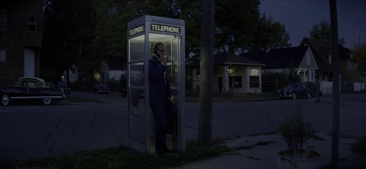 No Sudden Move (2021) movie still: wide — A quiet suburban street at night contains a single illuminated glass telephone booth. A man in a dark suit…