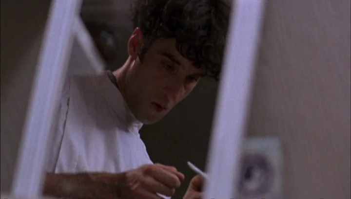 Highway 61 (1991) movie still: closeup — A man stands in a dimly lit bathroom while looking down at his hands. White bathroom fixtures and wall…