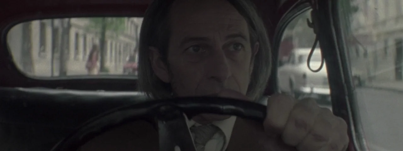 Post Mortem (2010) movie still: closeup — A man drives a vintage car through a blurry, overcast urban street environment. His focused face is framed…