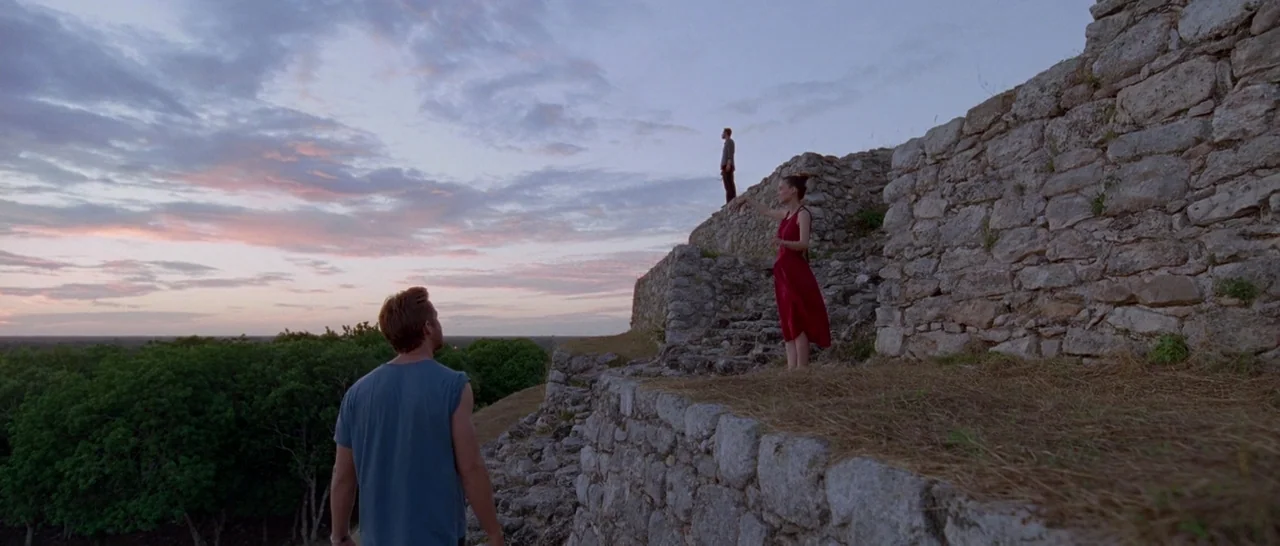 Song to Song (2017) movie still: wide — Ancient stone structures stand amidst a grassy landscape under a dusk sky. A man approaches a woman in a red…