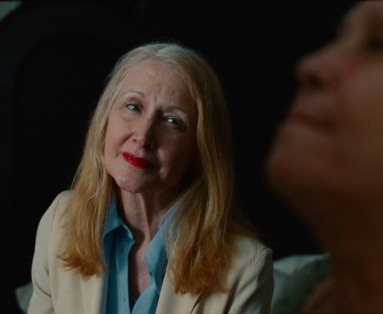 Monica (2022) movie still: medium — A woman sits in a dark, indistinct room. She glances towards an out-of-focus person nearby.
