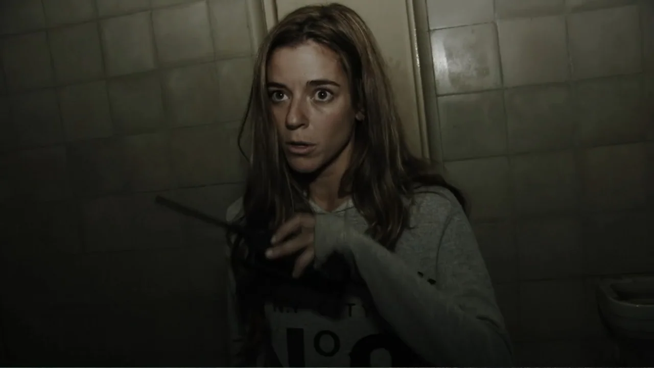 Lost (2010) movie still: medium — A dimly lit room with plain, square off-white wall tiles and a porcelain fixture. A woman stands in front of the…