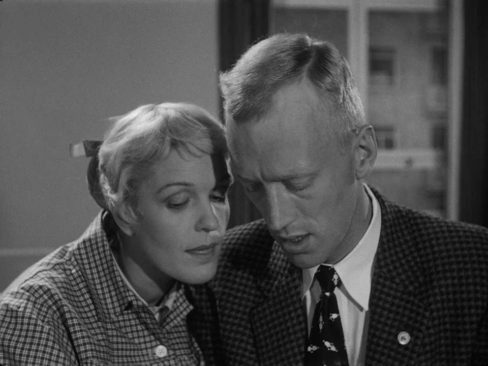 Brink of Life (1958) movie still: medium — Two individuals stand closely together inside a room with a window in the background. A man and a woman…