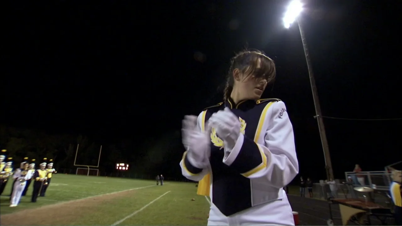 Life According to Sam (2013) movie still: medium — A nighttime view of a grassy sports field under a bright stadium light. A female marching band…