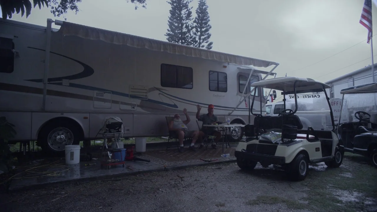 Florida Man (2015) movie still: wide — An RV park on an overcast day with rain visible in the air. Two older men sit in chairs beneath an RV awning…