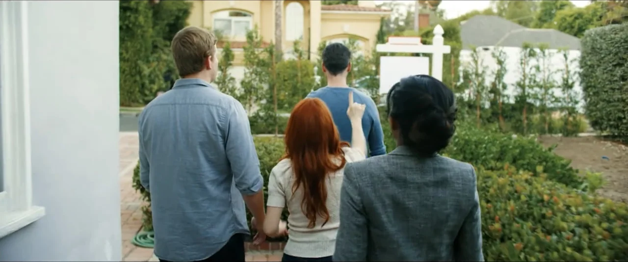 Gatlopp: Hell of a Game (2022) movie still: fullbody — A residential front yard features a large house and a real estate sign. Three people stand…