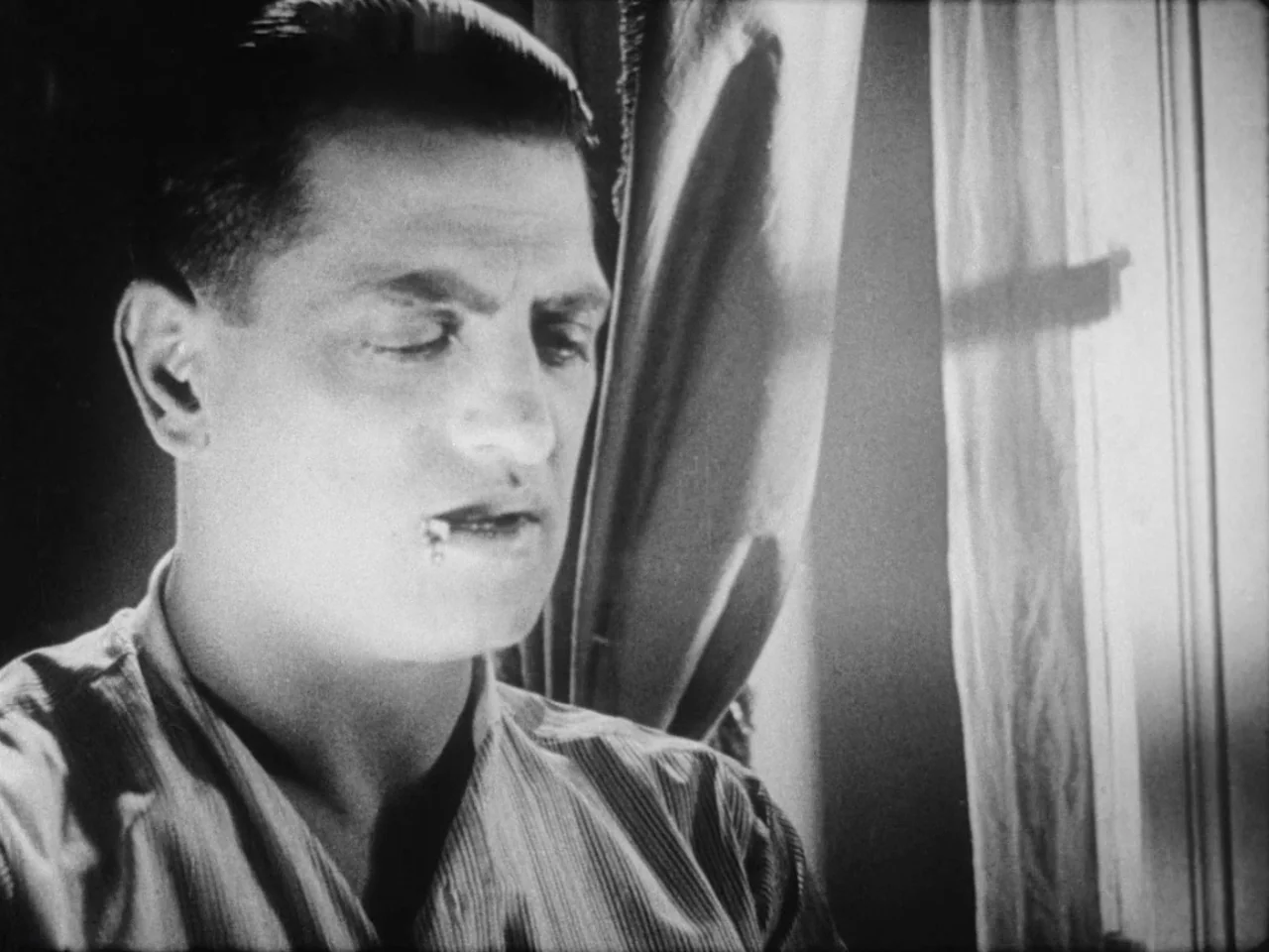 Un Chien Andalou (1929) movie still: closeup — A man stands indoors near a window with sheer curtains. He keeps his eyes closed while facing toward…